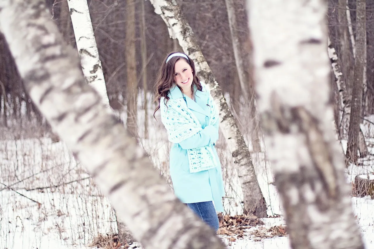 Femme portant un manteau d'hiver bleu clair, debout dans une forêt de bouleaux enneigée, lumière du jour